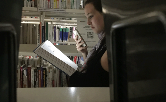 Jeune fille lisant dans la pénombre, entre deux rayons de livres, dans une bibliothèque ; elle éclaire son livre avec son smartphone
